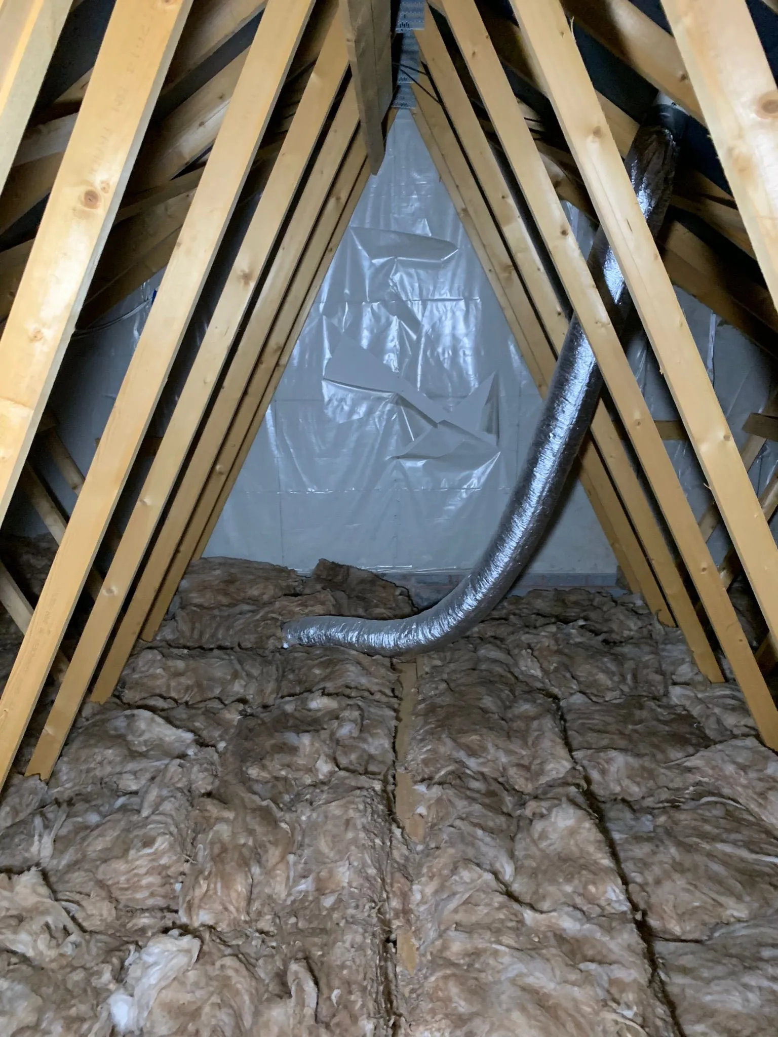 Attic space with wood beams, insulation material on the floor, and a flexible ducting pipe visible.