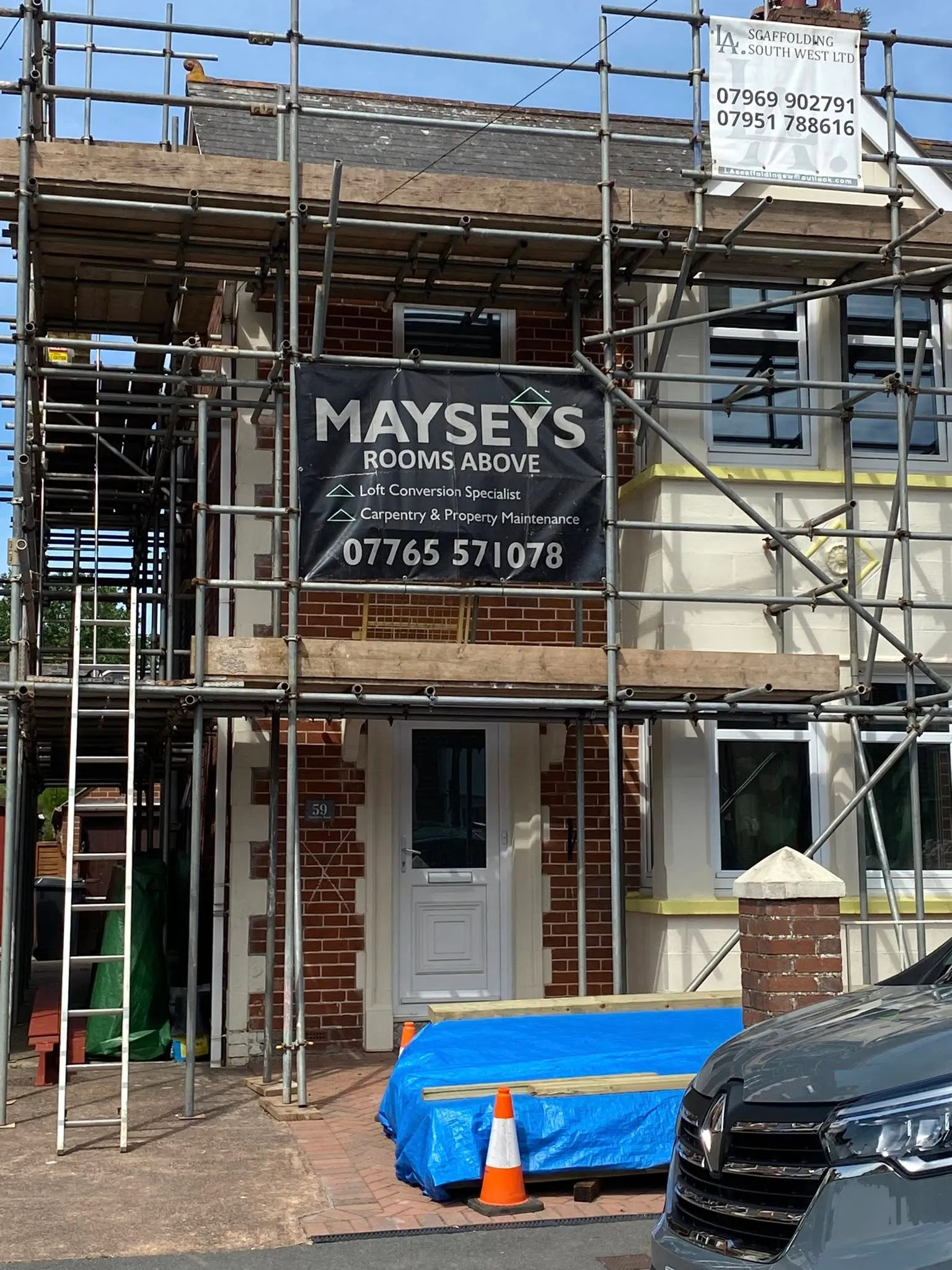 Home under renovation with scaffolding, a sign for Maysey's loft conversion, and a blue tarp covering wooden materials.