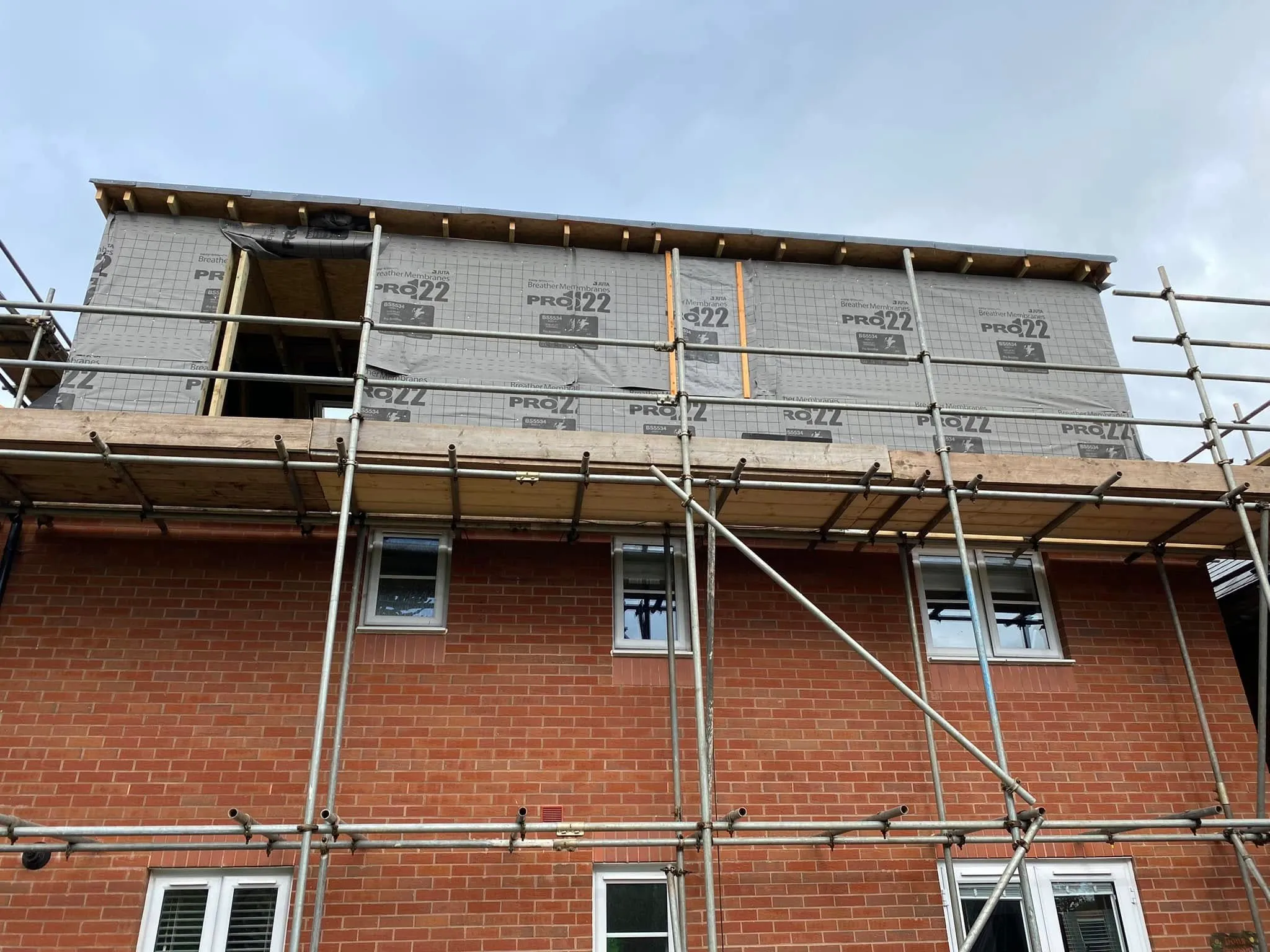 Construction site featuring scaffolding around a brick house with new materials, including insulation, on the upper level.