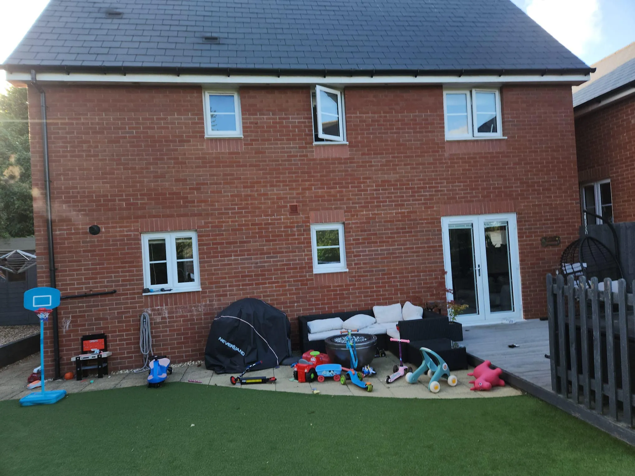 Backyard view of a brick house with two floors, toys scattered on artificial grass, a covered seating area, and a decorative fence.