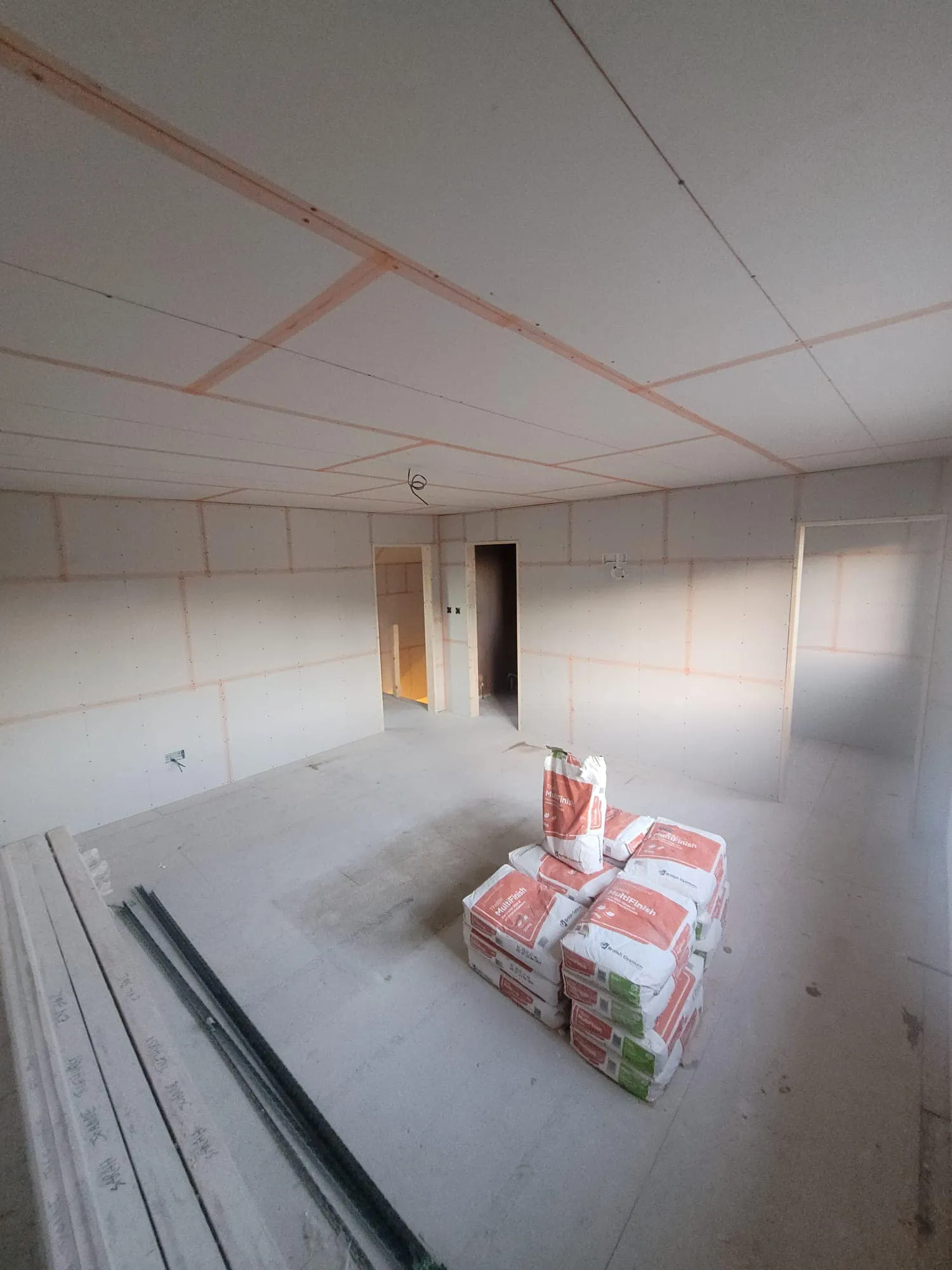 Interior of a partially constructed room with drywall, wooden beams, and stacked bags of building materials. Light coming from a doorway.