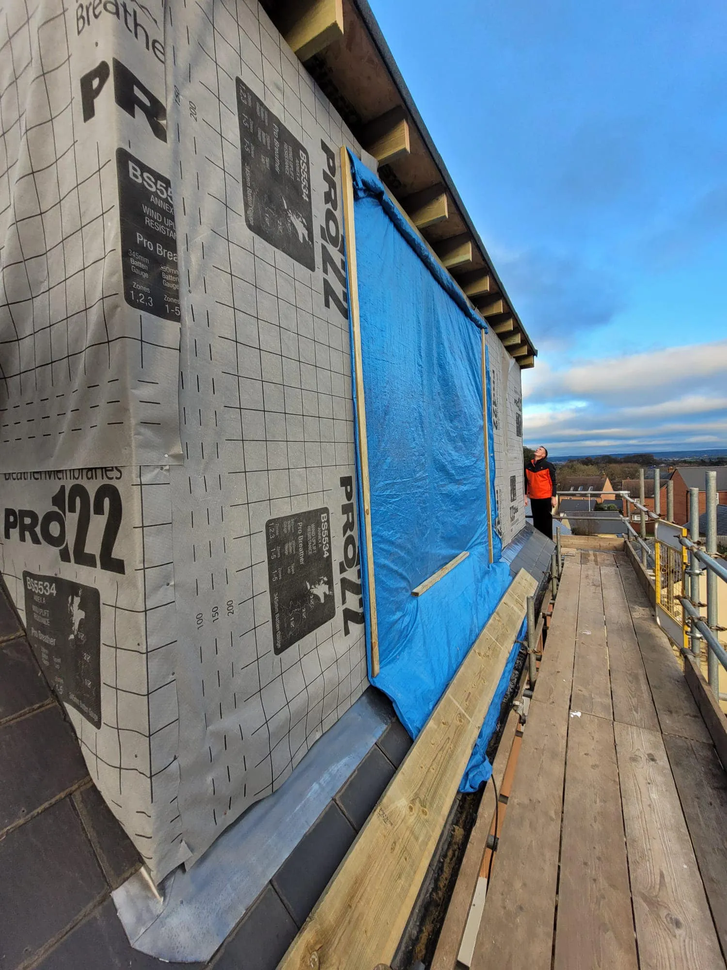 Construction site photo showing a roof with breathable membrane, blue tarp, and wooden scaffolding, under a partly cloudy sky.