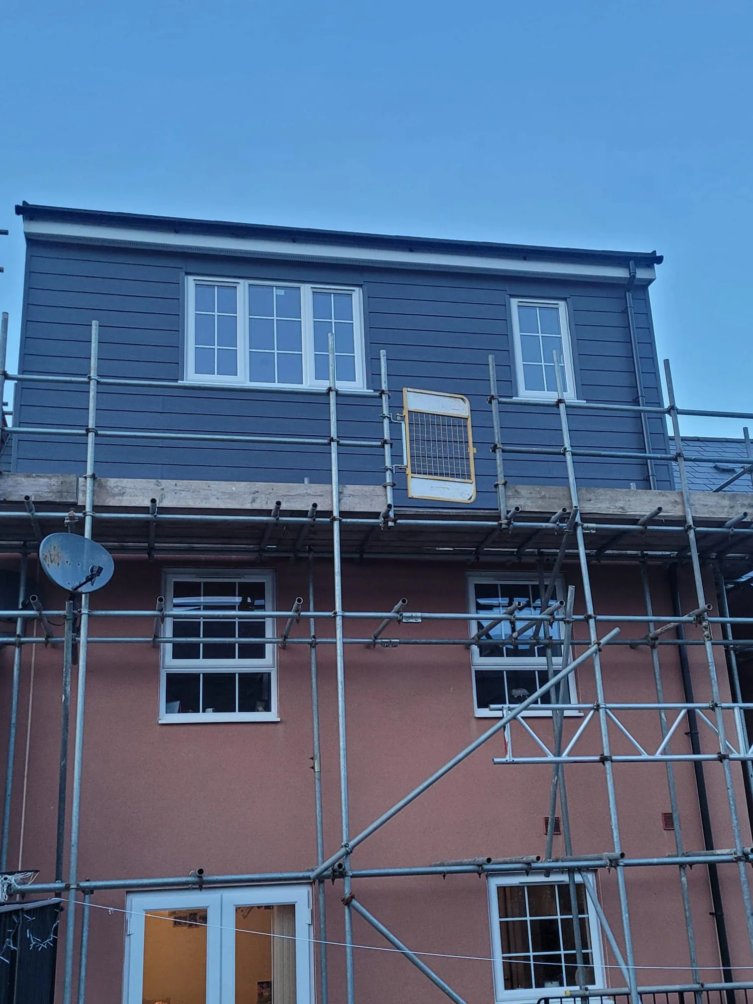 A building with scaffolding extends to a newly constructed upper section, featuring gray siding and multiple windows against a blue sky.