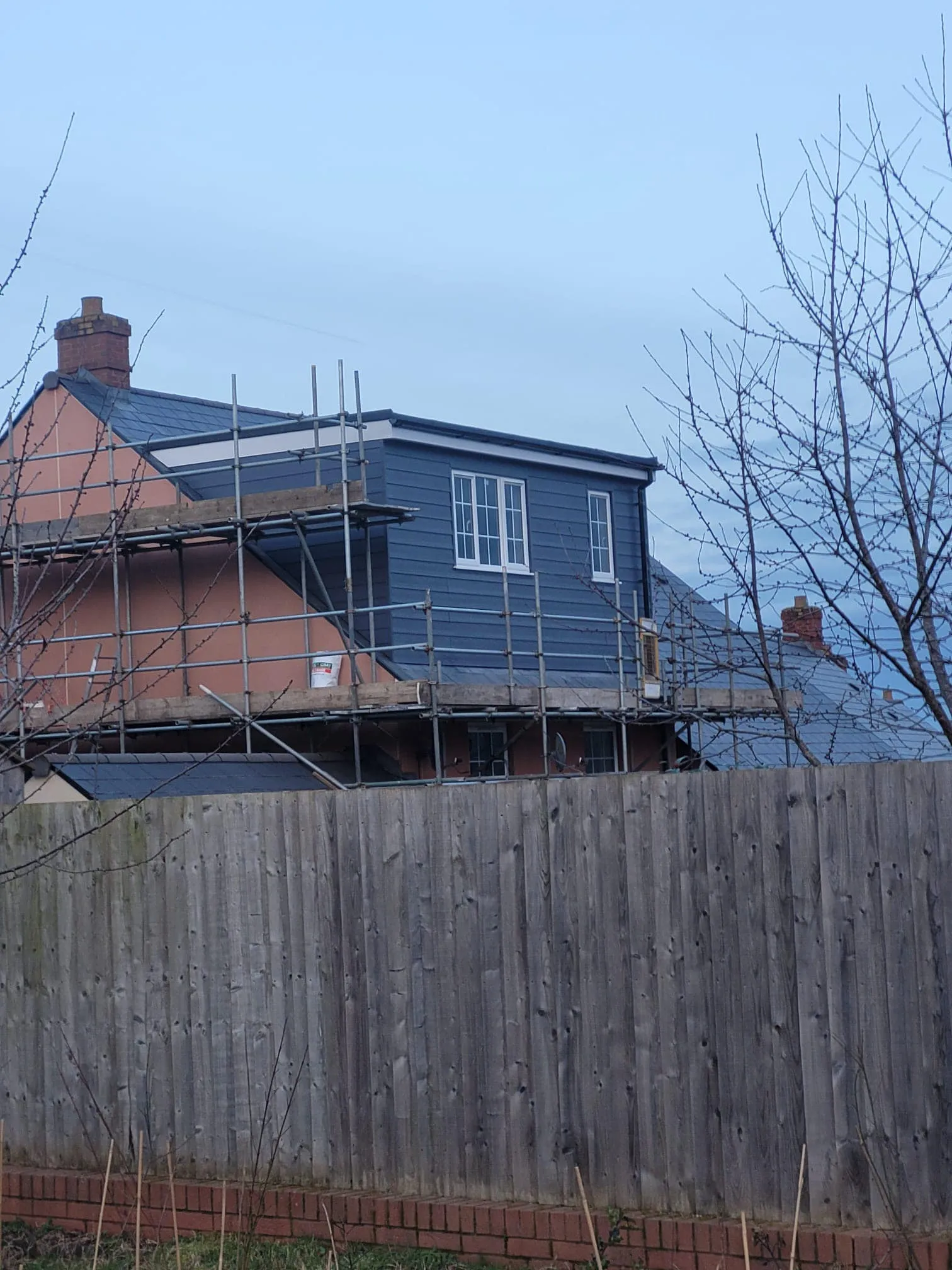 A house under construction with scaffolding, featuring a painted blue upper section and a peach lower section, set behind a wooden fence.
