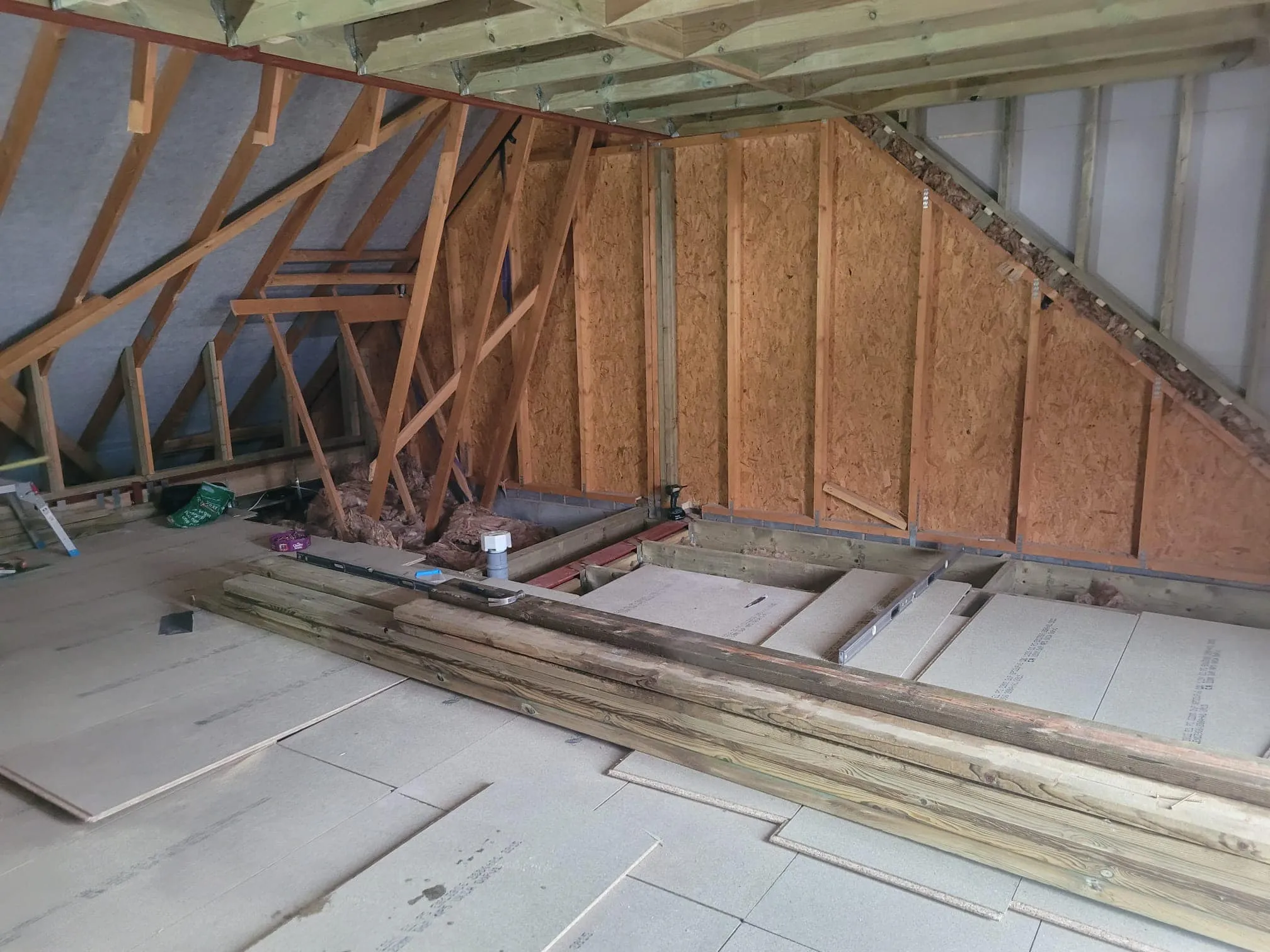 An unfinished attic space featuring exposed wooden beams, insulation material, and stacked wooden planks on the floor.