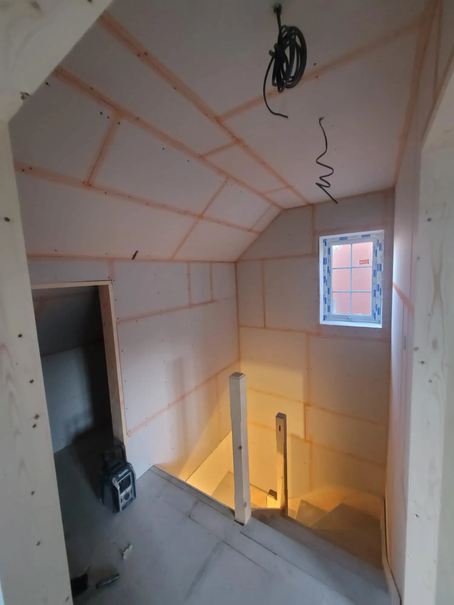Interior view of a partially constructed room with drywall and exposed beams, showing a staircase and a window.