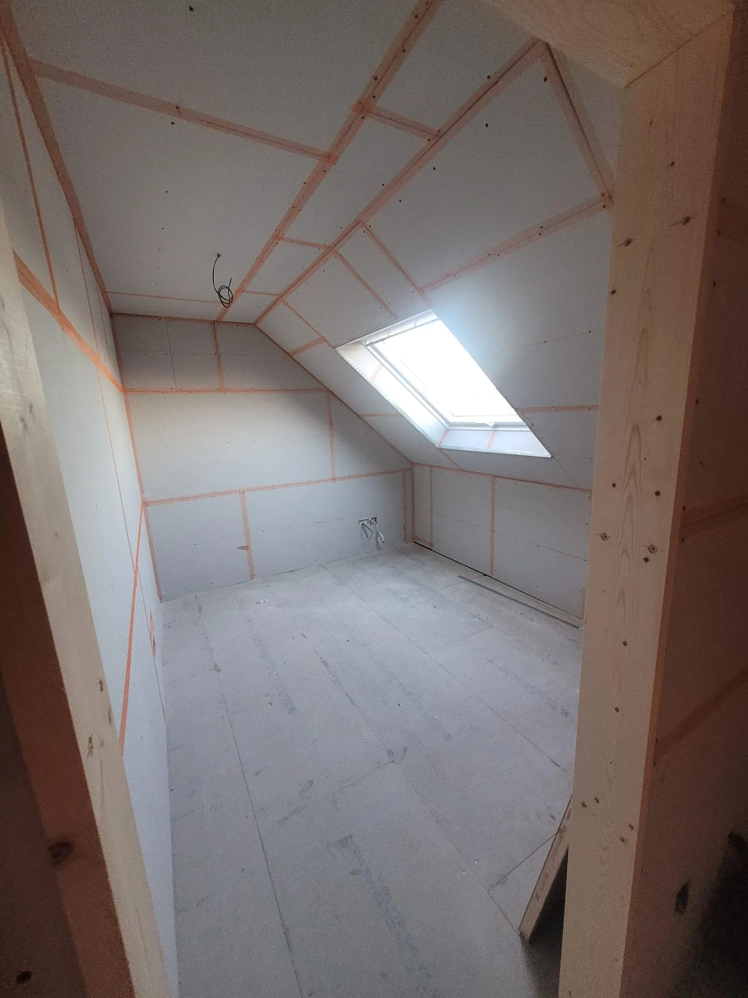 A partially finished room with slanted walls, a skylight, and exposed drywall framing. The floor is bare wood.
