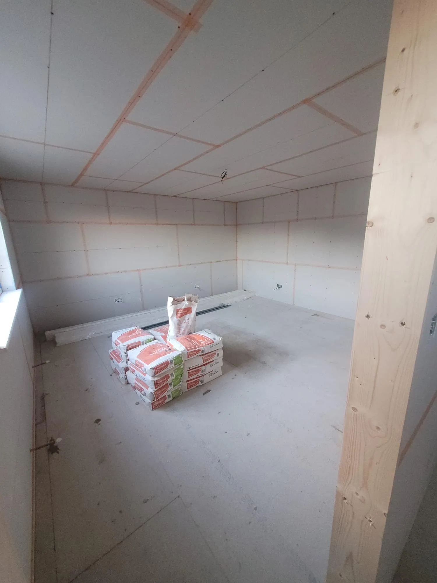 An unfinished room with drywall on walls and ceiling, containing a stack of construction materials on the floor.