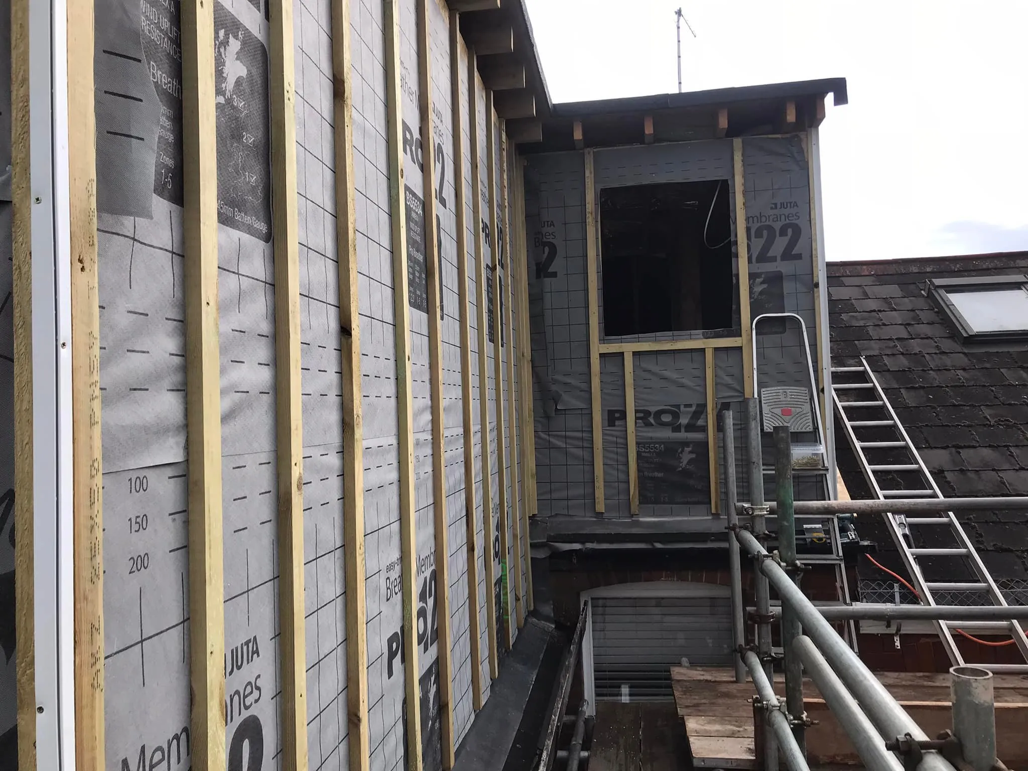 Construction site showing wooden framing with weather-resistant membrane, scaffolding, and a ladder against a roof.