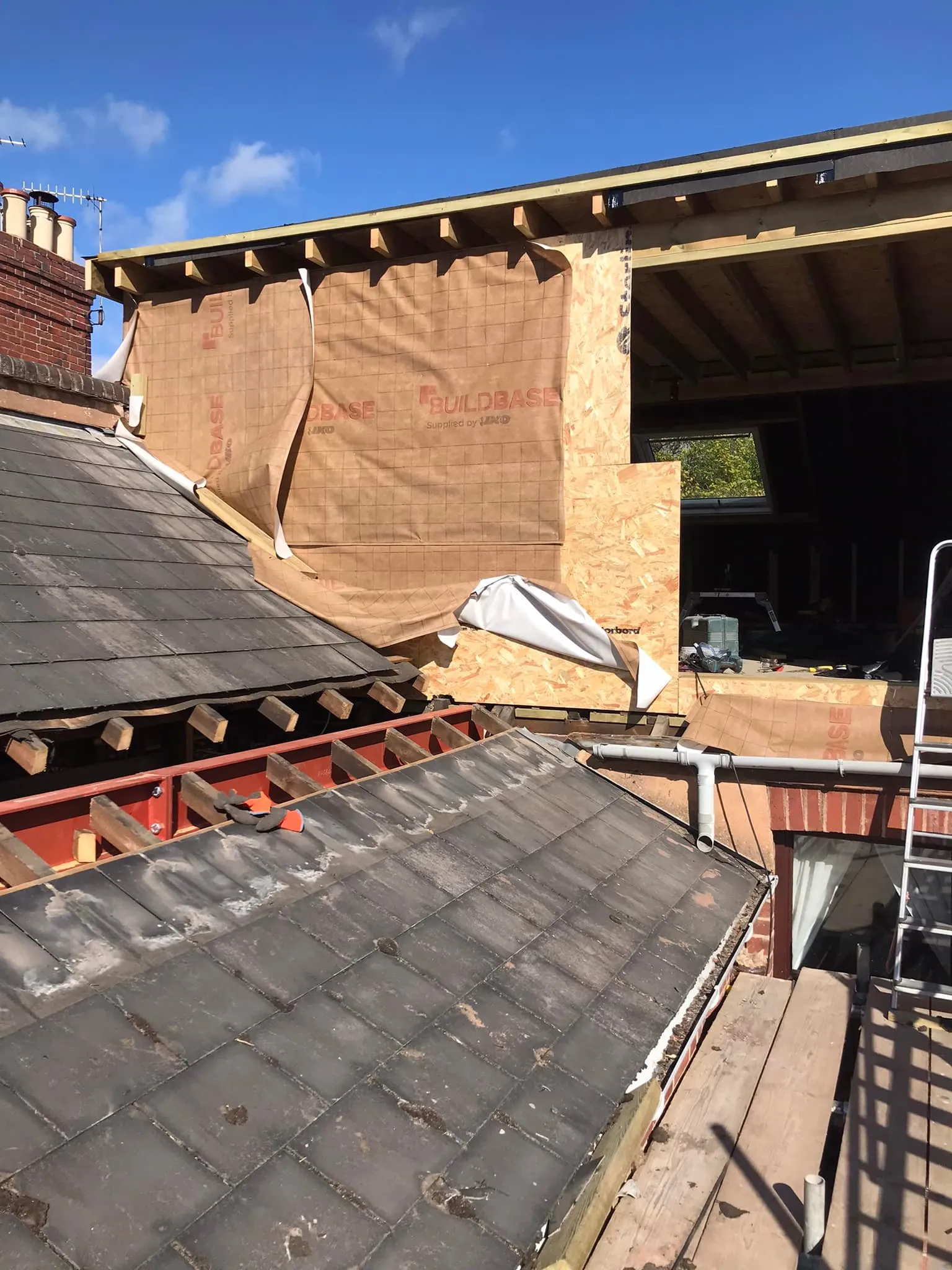 Construction scene showing a roof extension with exposed beams, construction materials, and a partially covered area. Bright blue sky above.