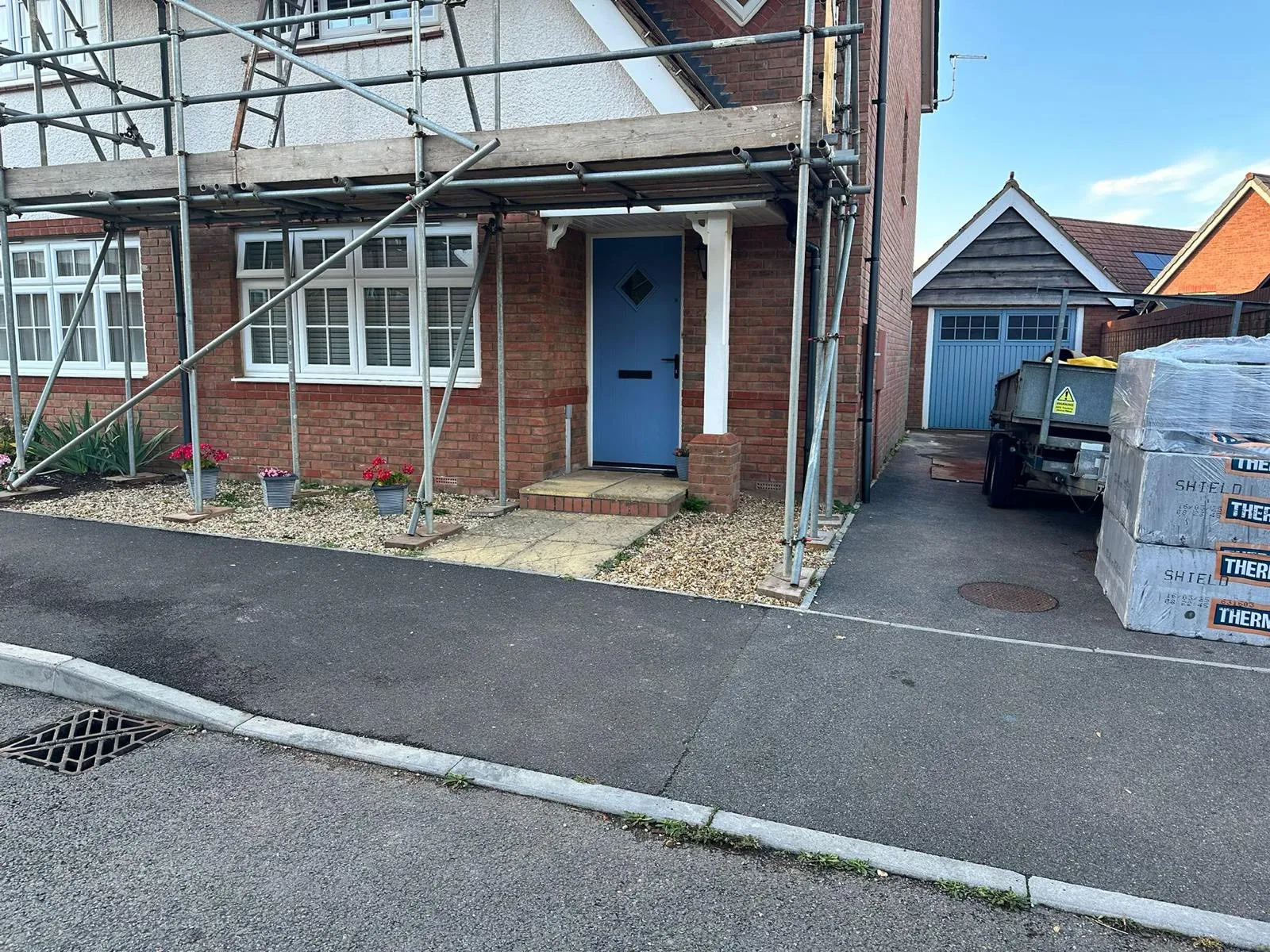 A house with scaffolding in front, a blue door, flower pots, and a parked trailer on the side. Road is asphalt with a drain.