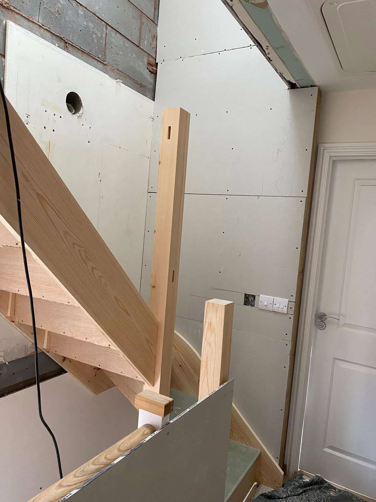 Construction near stairs, featuring wooden stair railings, unfinished drywalls, and electrical outlets. Tools and debris on the floor.