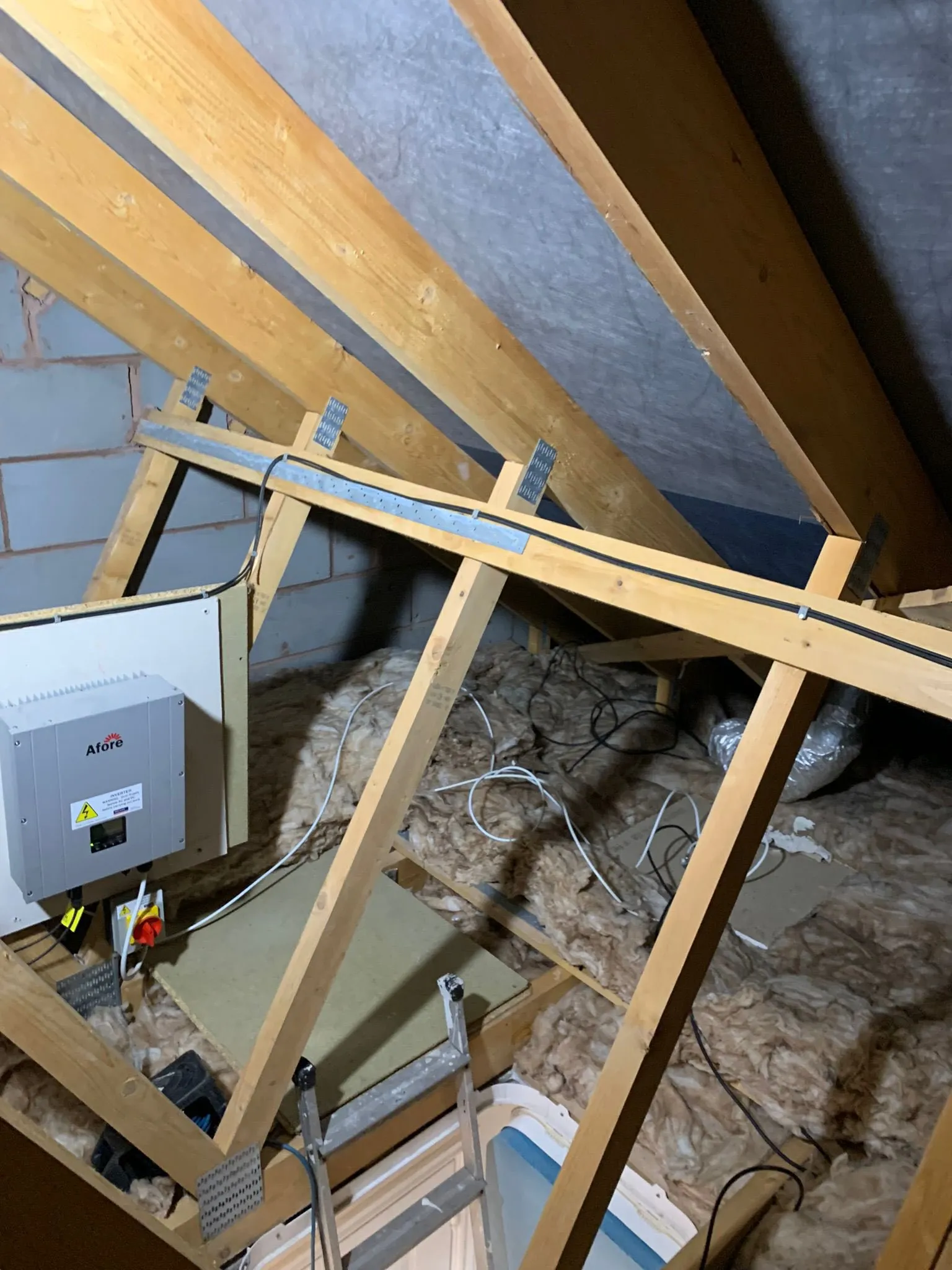 Attic interior with wooden beams, insulation, a solar inverter, and wires. A metal ladder is visible on the flooring.
