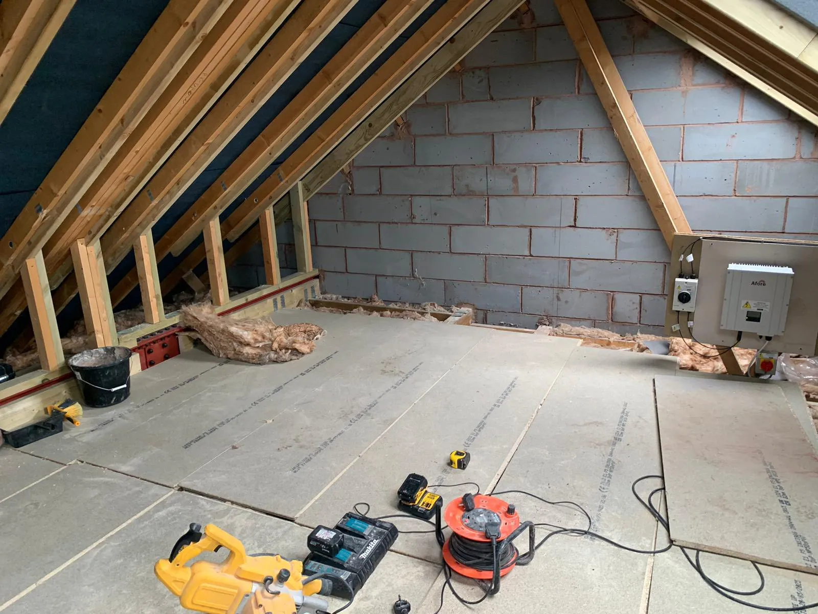 Attic space under construction with wooden beams, insulation, tools, and a wall-mounted device, featuring a concrete block wall.